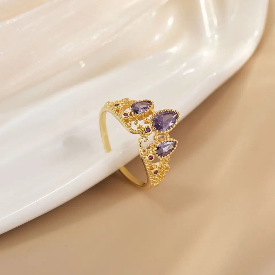 Anillo dorado con diseño de corona y piedras moradas sobre una superficie de nácar