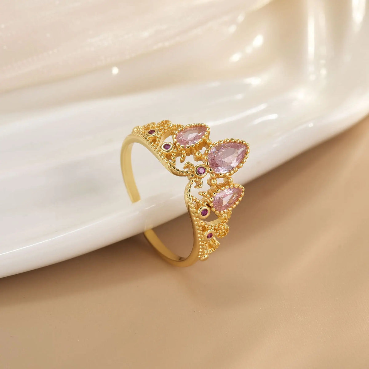 Anillo dorado con diseño de corona y piedras rosadas sobre una superficie de nácar